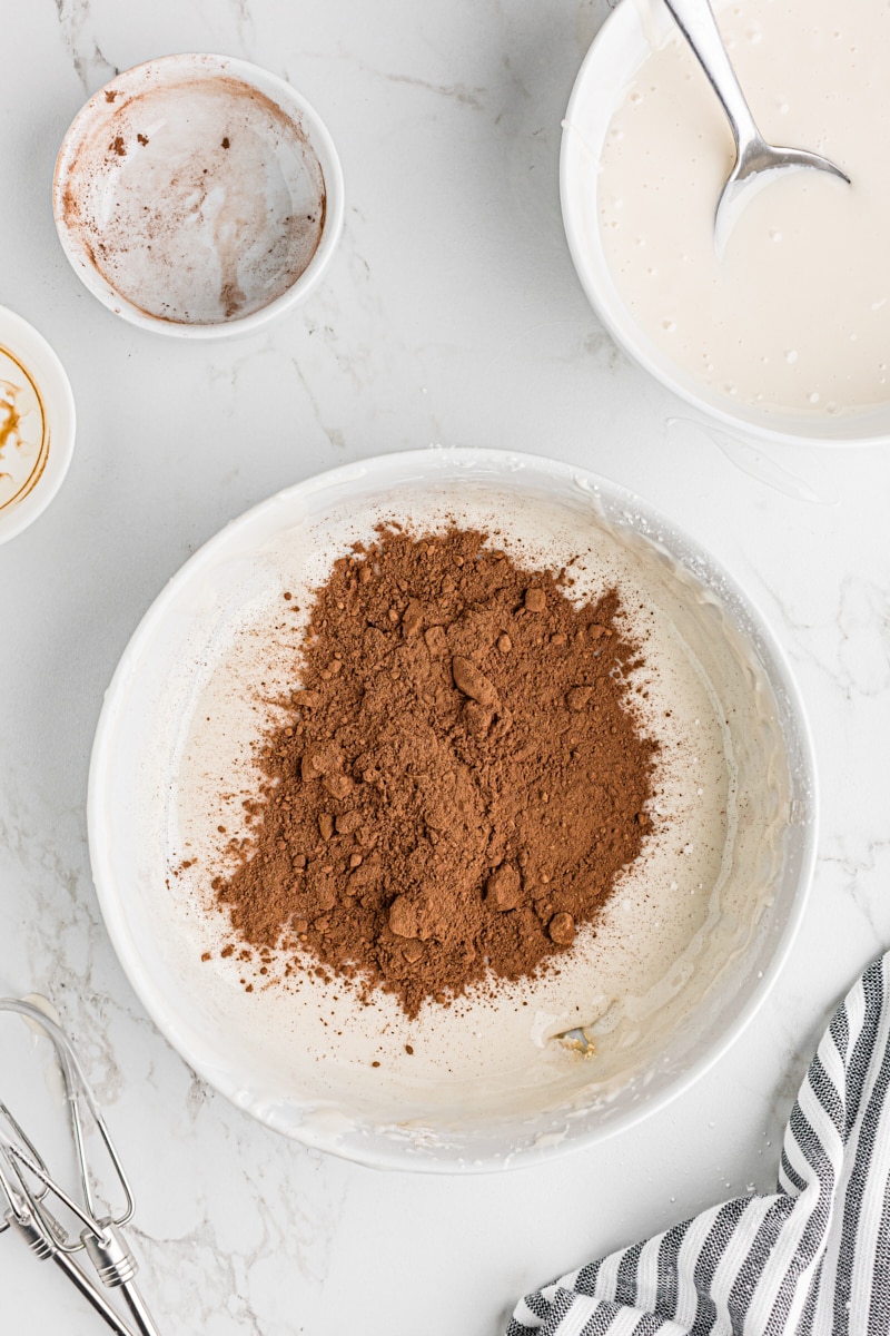 Cocoa powder being added to a portion of the vanilla icing in a white bowl to create chocolate icing.