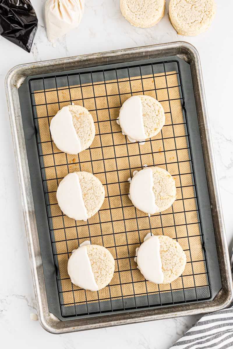 Black and white cookies partially iced with vanilla frosting, resting on a wire rack over a baking sheet.