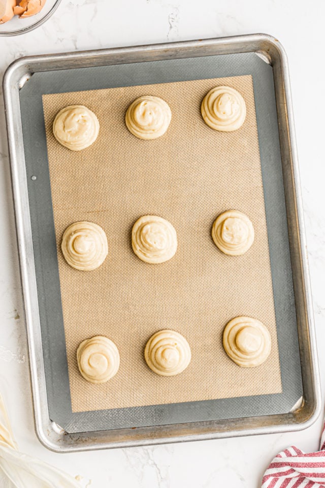overhead view of piped pâte à choux on a baking sheet lined with a silicone baking mat