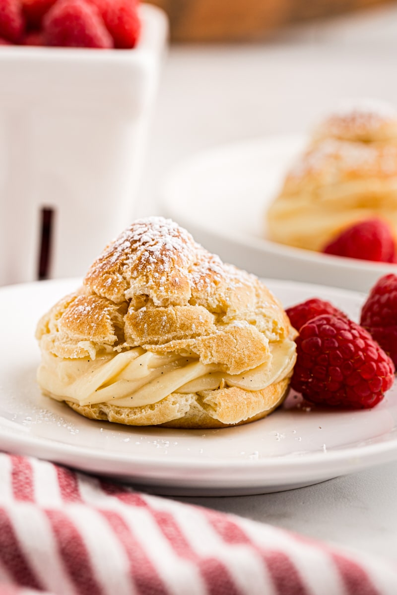 a cream puff and raspberries on a white plate with a container of raspberries and another cream puff in the background