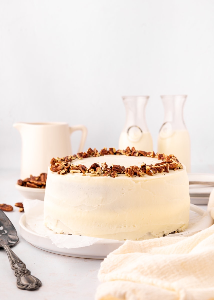 a carrot cake on a white cake plate with two carafes of milk, a small white pitcher, and a bowl of pecans in the background