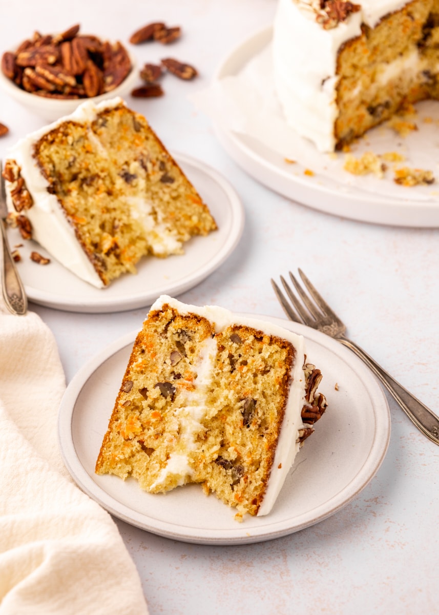 two slices of carrot cake served on light beige plates with the remaining cake and a bowl of pecans in the background