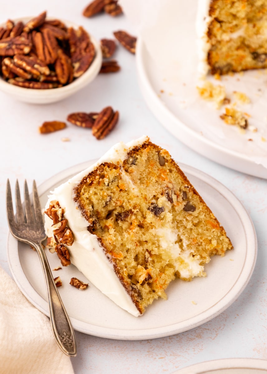 a slice of carrot cake on a light beige plate with more cake and a bowl of pecans in the background
