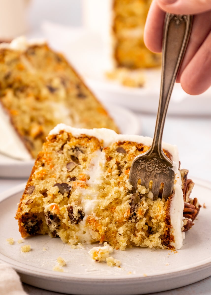 a fork cutting into a slice of carrot cake; another slice of cake and the remaining cake are in the background