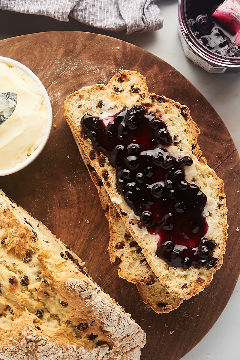 overhead view of a slice of Irish soda bread with butter and blueberry preserves spread onto it; more bread, butter, and preserves alongside