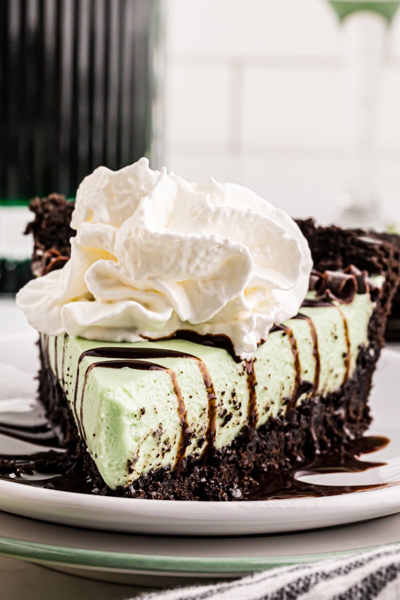 a slice of grasshopper pie on a white plate topped with chocolate curls, chocolate syrup, and whipped cream