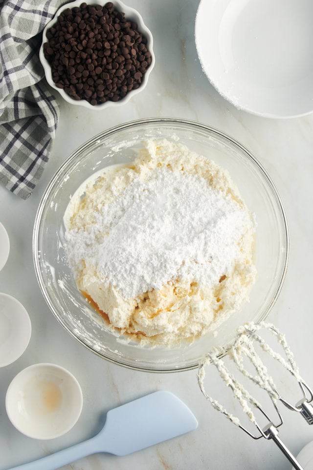 overhead view of confectioners' sugar, vanilla extract, and almond extract added to combined mascarpone and ricotta cheeses in a glass mixing bowl