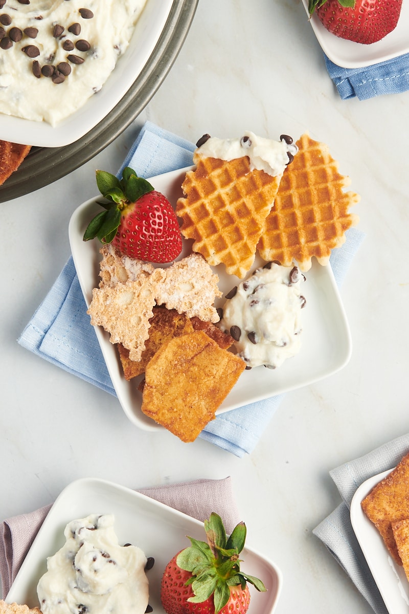 cannoli dip served on a square white plate with waffle cookies, strawberry, and pita chips; more plates surrounding