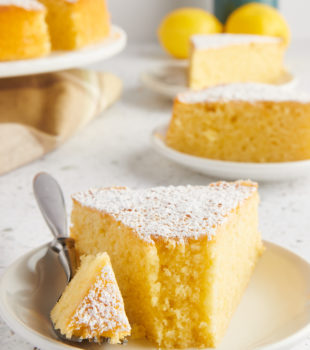 a slice of olive oil cake on a white plate with a bite on a fork; more cake and lemons in the background