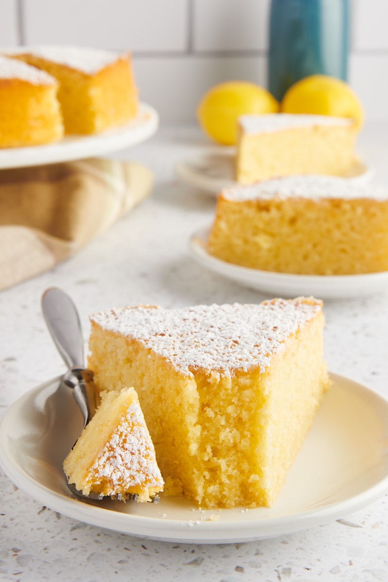 a slice of olive oil cake on a white plate with a bite on a fork; more cake and lemons in the background