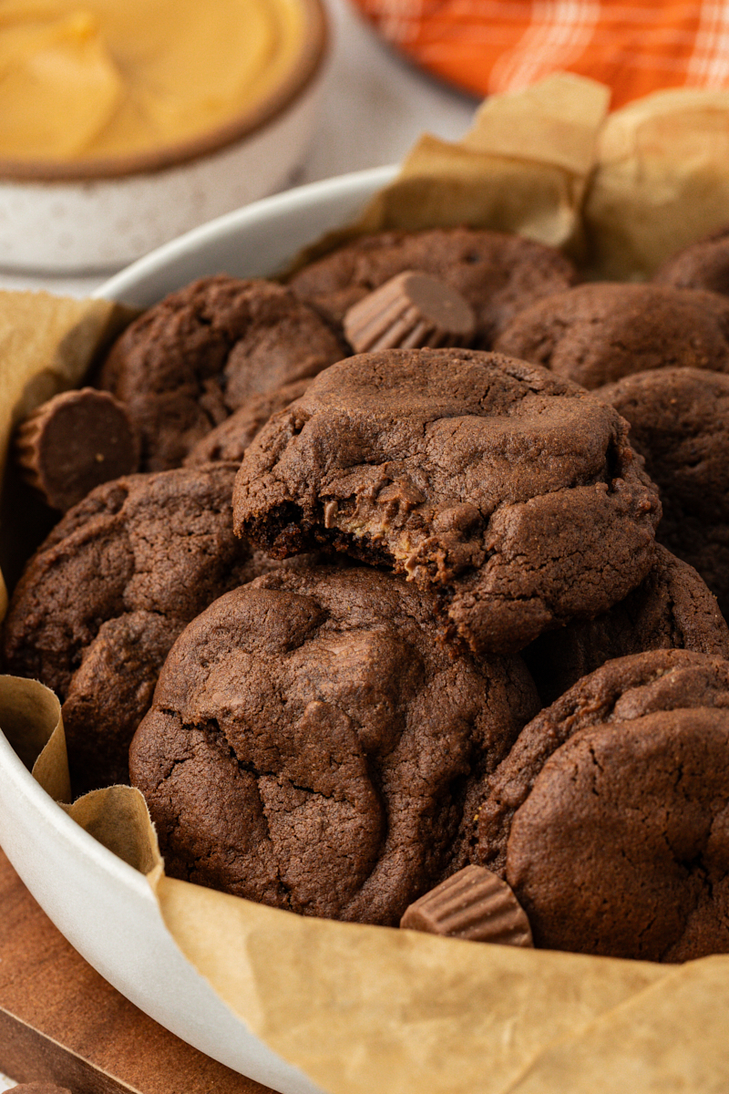chocolate peanut butter cookies piled in a large white bowl with the cookie on top missing a bite to show the peanut butter cup filling