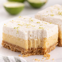 a key lime pie bar with a bite missing on a white plate with another bar and limes in the background