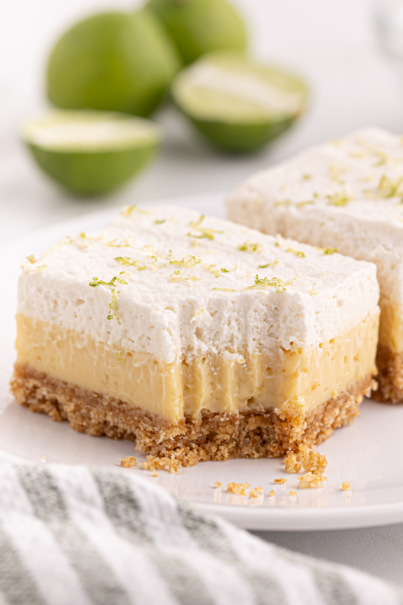 a key lime pie bar with a bite missing on a white plate with another bar and limes in the background