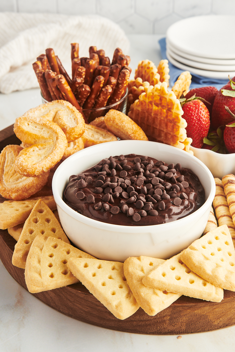 a wooden board with a bowl of brownie batter dip and an assortment of cookies, berries, and pretzels for dipping