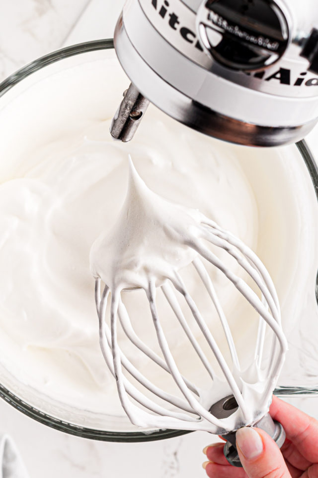 the whisk of a stand mixer lifted out of a bowl of whipped egg whites to show stiff peaks