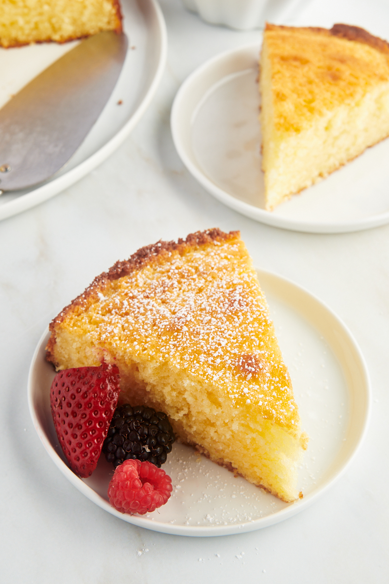a slice of lemon ricotta cake on a white plate with more cake in the background