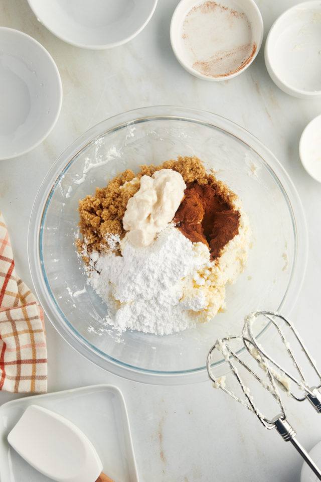 overhead view of confectioners' sugar, brown sugar, yogurt, cinnamon, and vanilla added to creamed butter and cream cheese in a glass mixing bowl