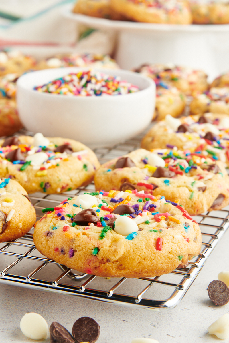 sprinkle chocolate chip cookies on a wire rack with a bowl of sprinkles and more cookies on a white cake stand in the background