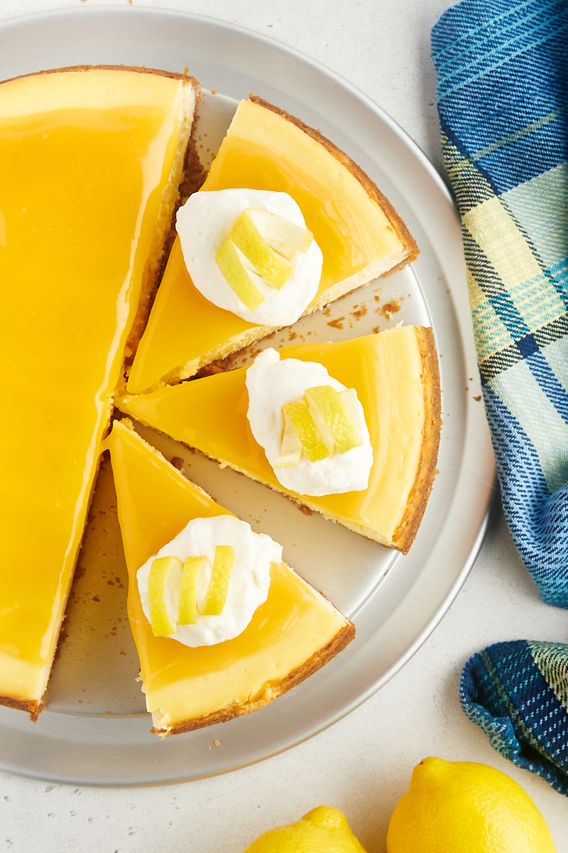 overhead view of partially sliced lemon cheesecake topped with lemon curd, whipped cream, and lemon curls