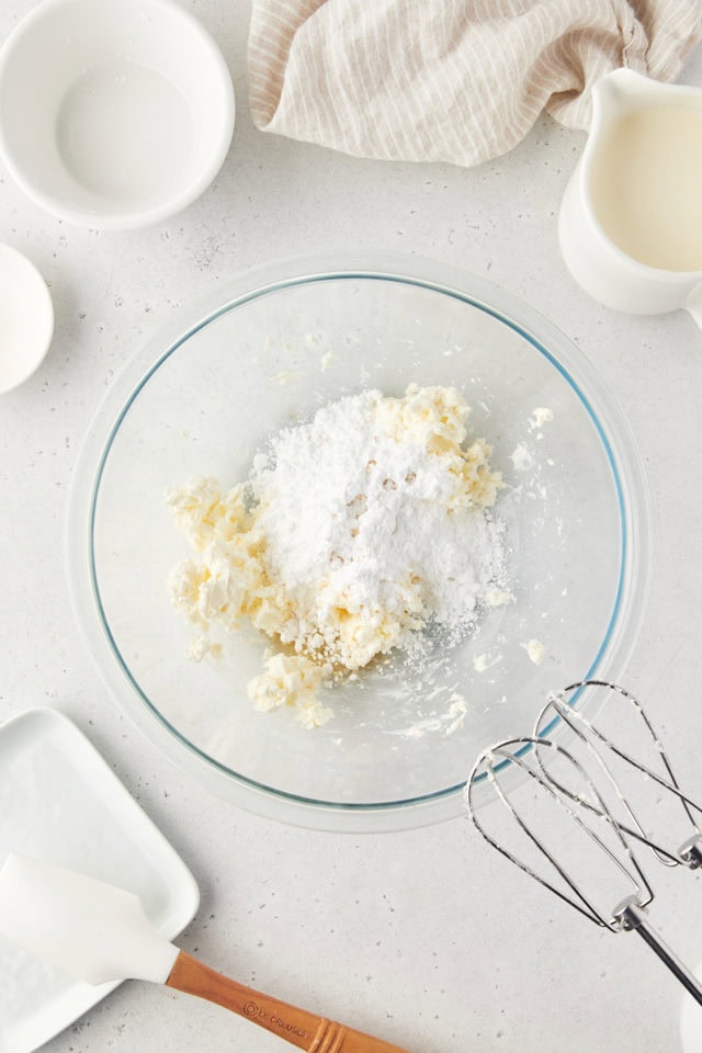 overhead view of confectioners' sugar and vanilla extract added to beaten cream cheese in a glass mixing bowl