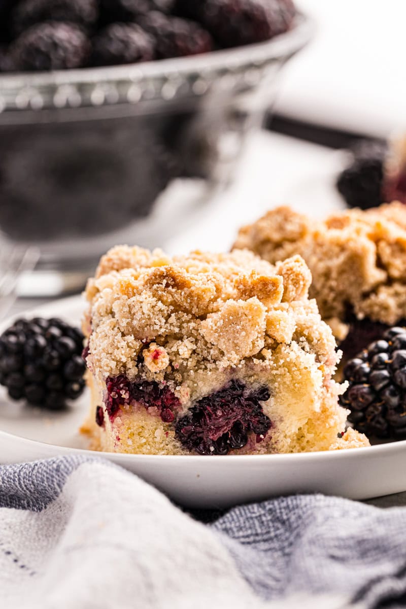 close-up view of a blackberry crumb bar on a white plate with a bowl of blackberries in the background
