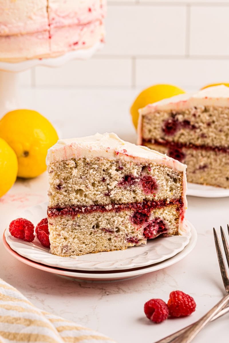 a slice of lemon raspberry cake on a white plate with more cake in the background