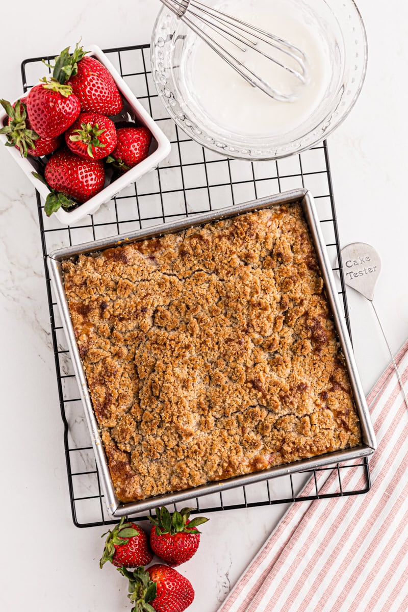 overhead view of freshly baked strawberry coffee cake on a wire rack with strawberries and freshly mixed glaze alongside