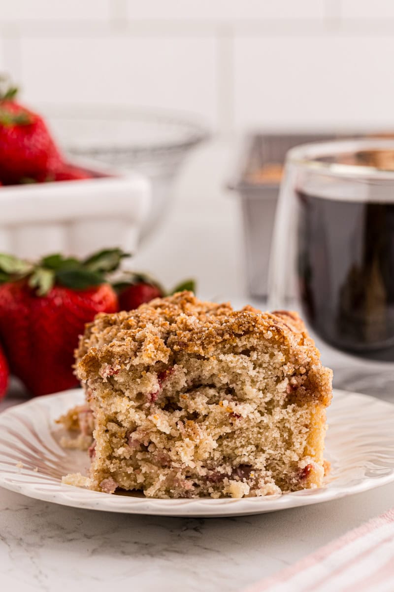 a slice of strawberry coffee cake on a white plate with a cup of coffee and fresh strawberries in the background