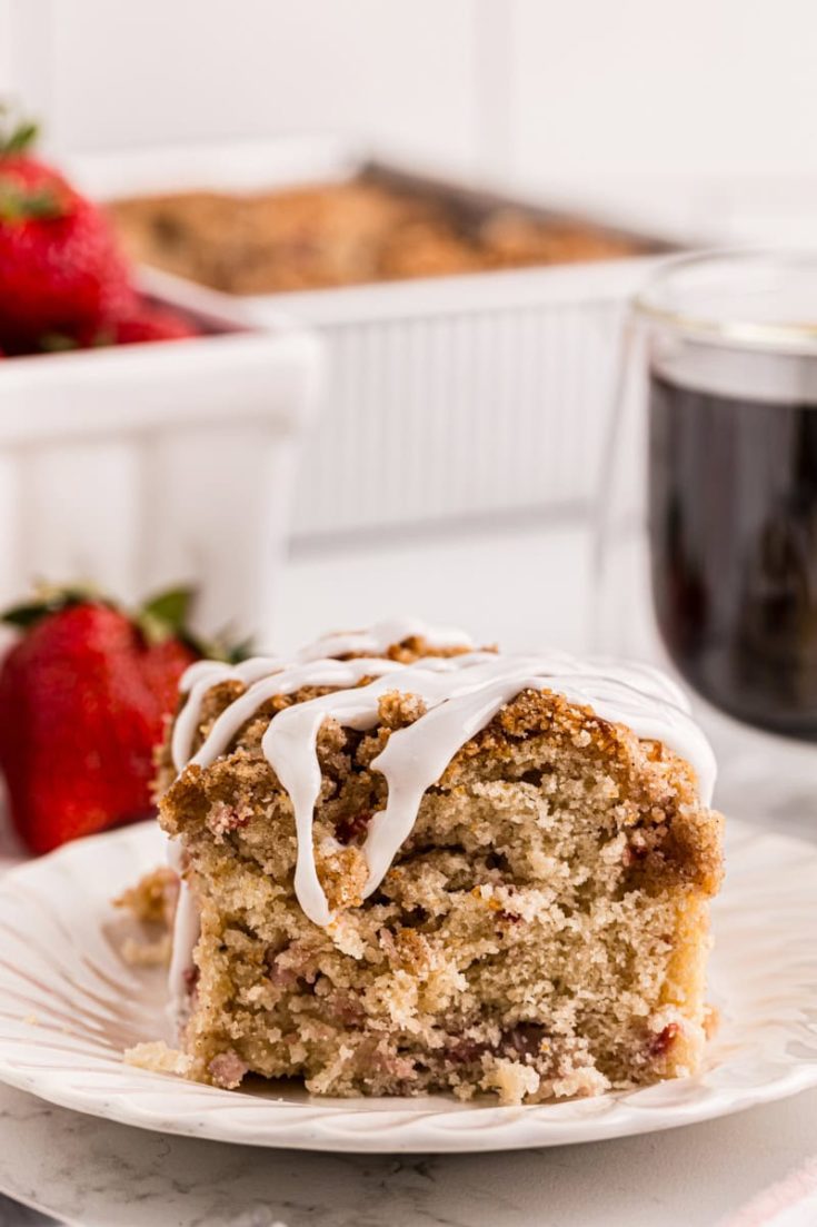 a slice of strawberry coffee cake on a white plate with berries and more cake in the background