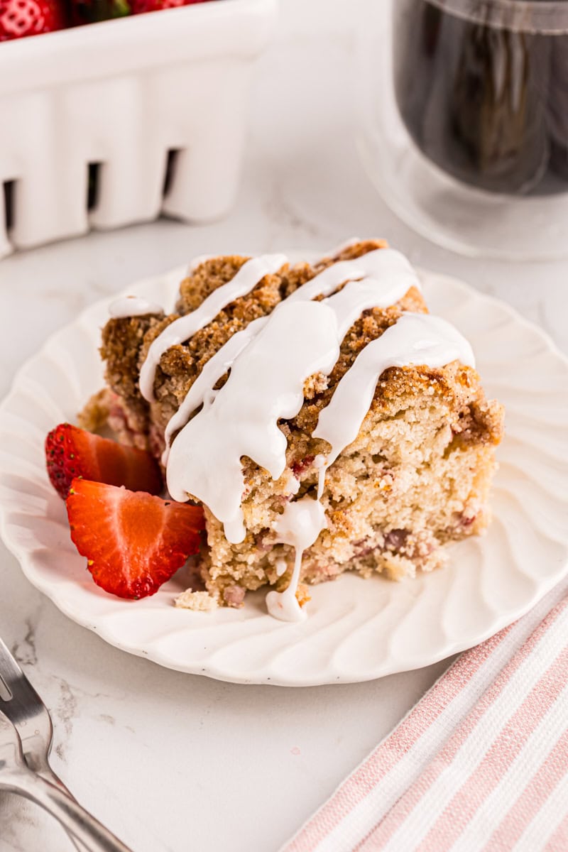 a slice of strawberry coffee cake topped with glaze and served on a white plate with sliced strawberries