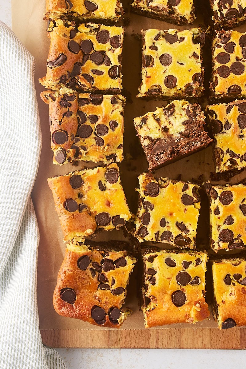 overhead view of chocolate chip cheesecake brownies on parchment paper with one brownie turned on its size to show the thickness of the layers