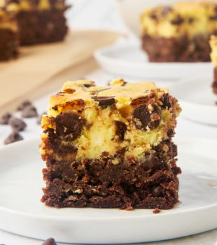 a chocolate chip cheesecake brownie on a white plate with more brownies in the background