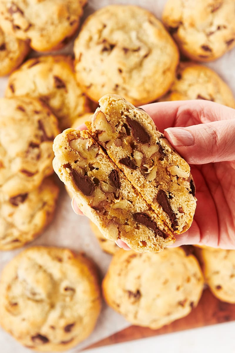 a hand holding two halves of a thick chocolate chip cookie with more cookies in the background