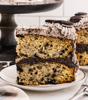 side view of a slice of cookies and cream cake on a white plate with more cake in the background