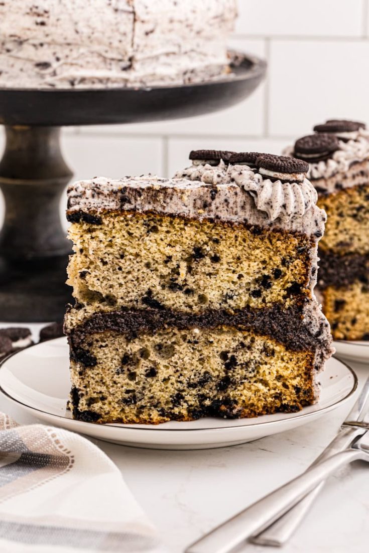 side view of a slice of cookies and cream cake on a white plate with more cake in the background