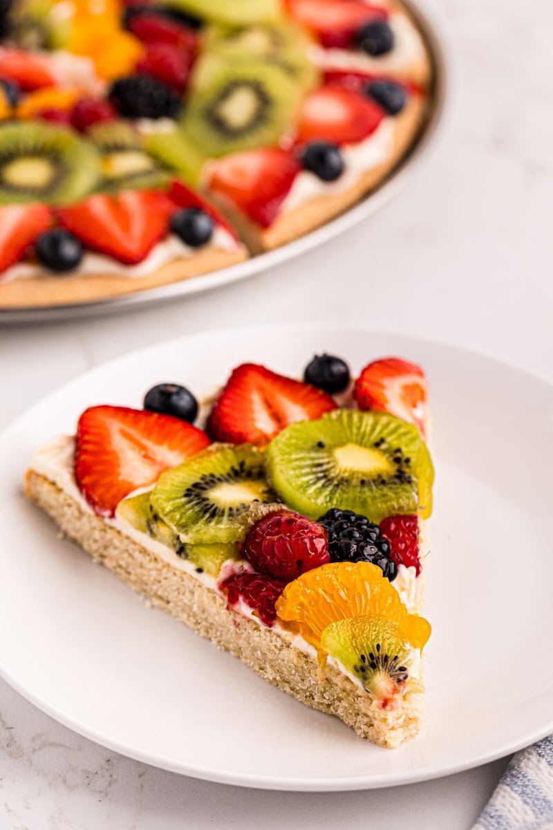 a slice of fruit pizza on a white plate with the remaining pizza in the background