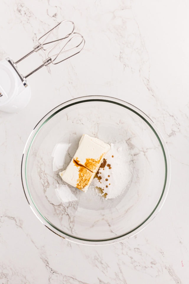 overhead view of cream cheese, sugar, and vanilla extract in a glass mixing bowl