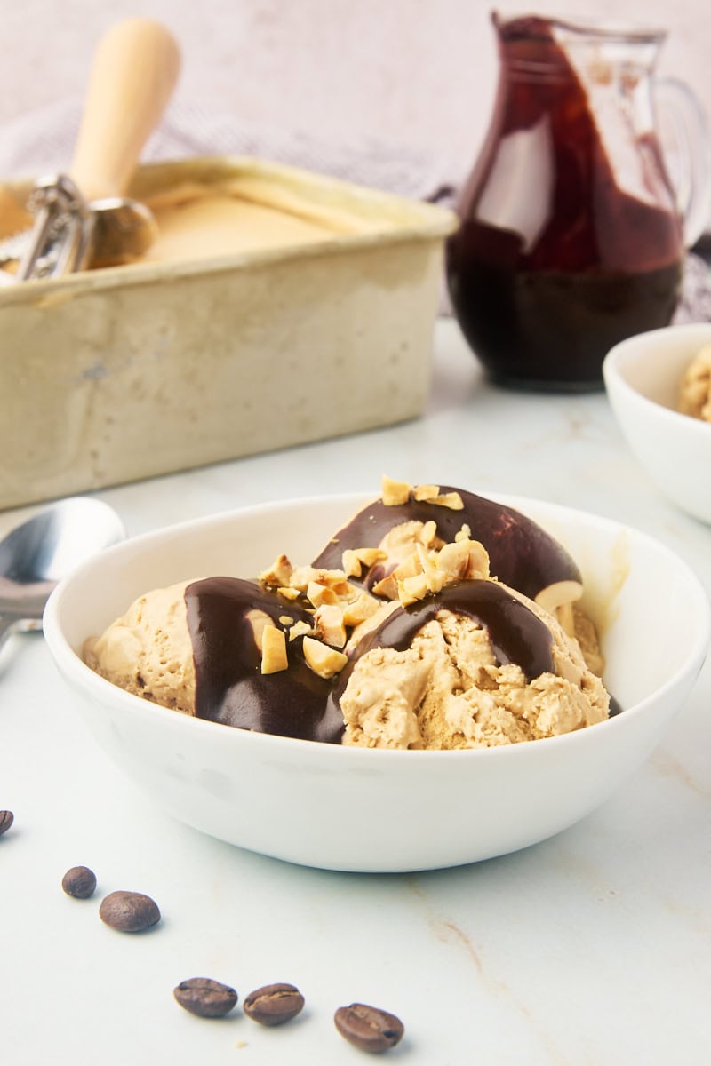 a bowl of coffee ice cream topped with hot fudge sauce, with more ice cream and fudge sauce in the background