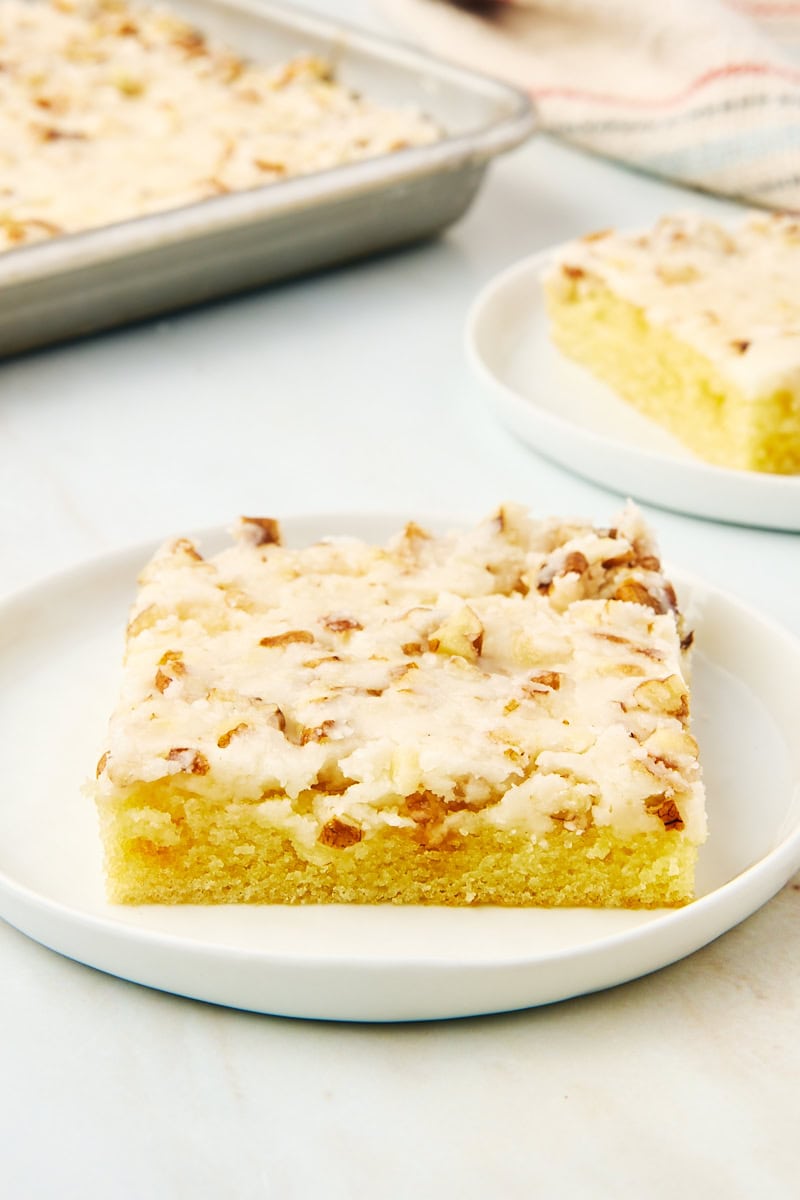 a slice of white Texas sheet cake on a white plate with more cake in the background