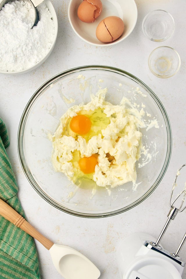 overhead view of eggs, vanilla, and salt added to beaten cream cheese in a glass mixing bowl