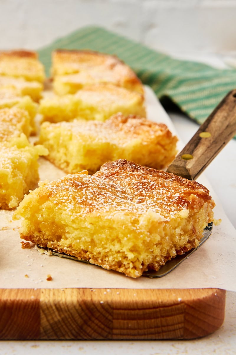 a slice of gooey butter cake on a serving spatula with more cake in the background