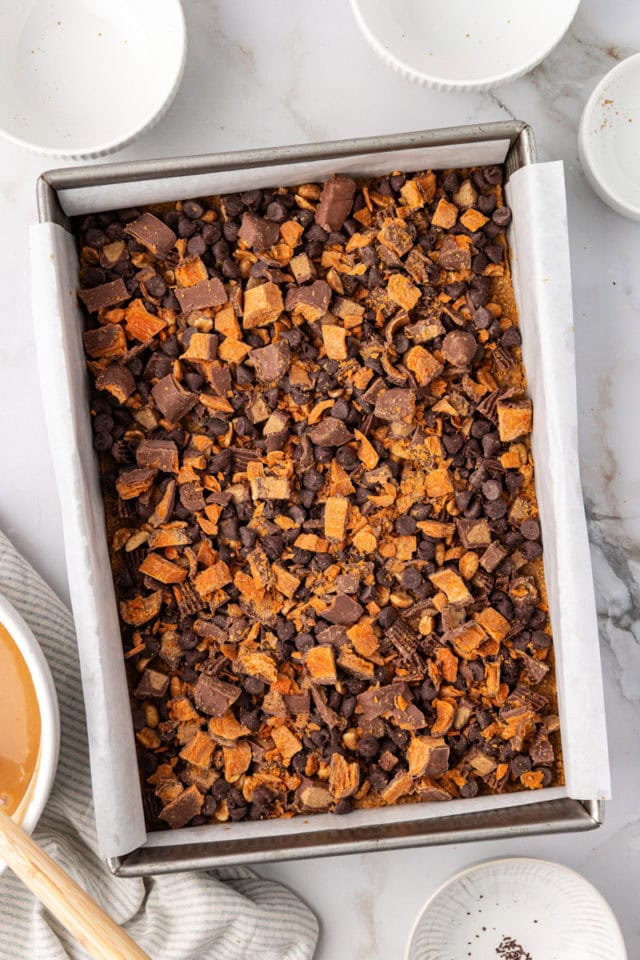 overhead view of candy bar pieces, chocolate chips, and peanuts on top of a Nutter Butter cookie crust in a 9x13 pan