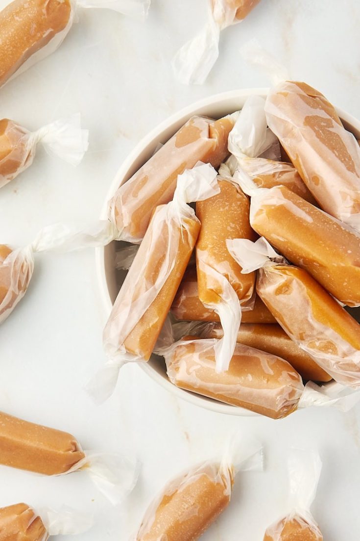 overhead view of homemade soft caramels in a white bowl with more caramels scattered around the bowl