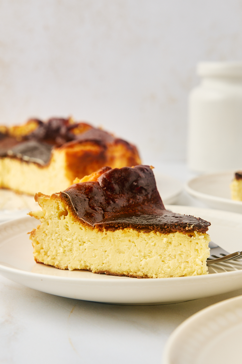 a slice of basque cheesecake on a white plate with more cheesecake in the background