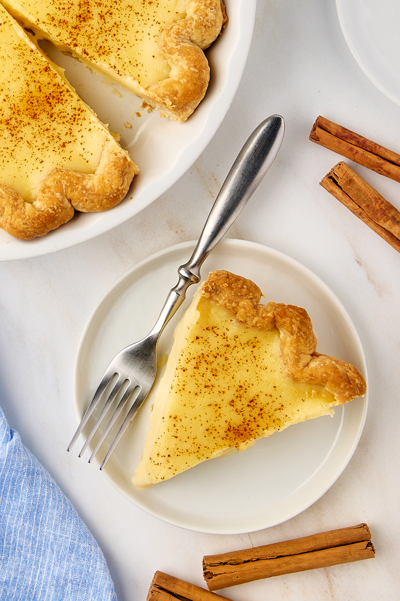 overhead view of a slice of sugar cream pie on a white plate beside the remaining pie in a pie plate