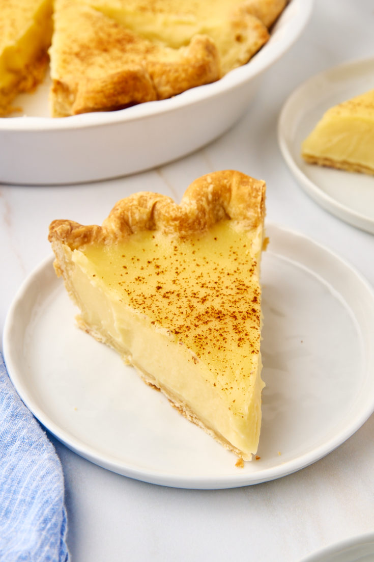 a slice of sugar cream pie on a white plate next to the remaining pie