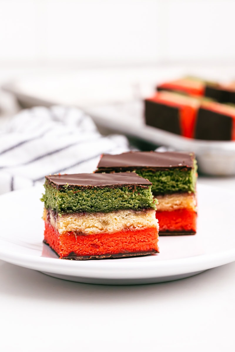 two Italian rainbow cookies on a white plate with more cookies in the background