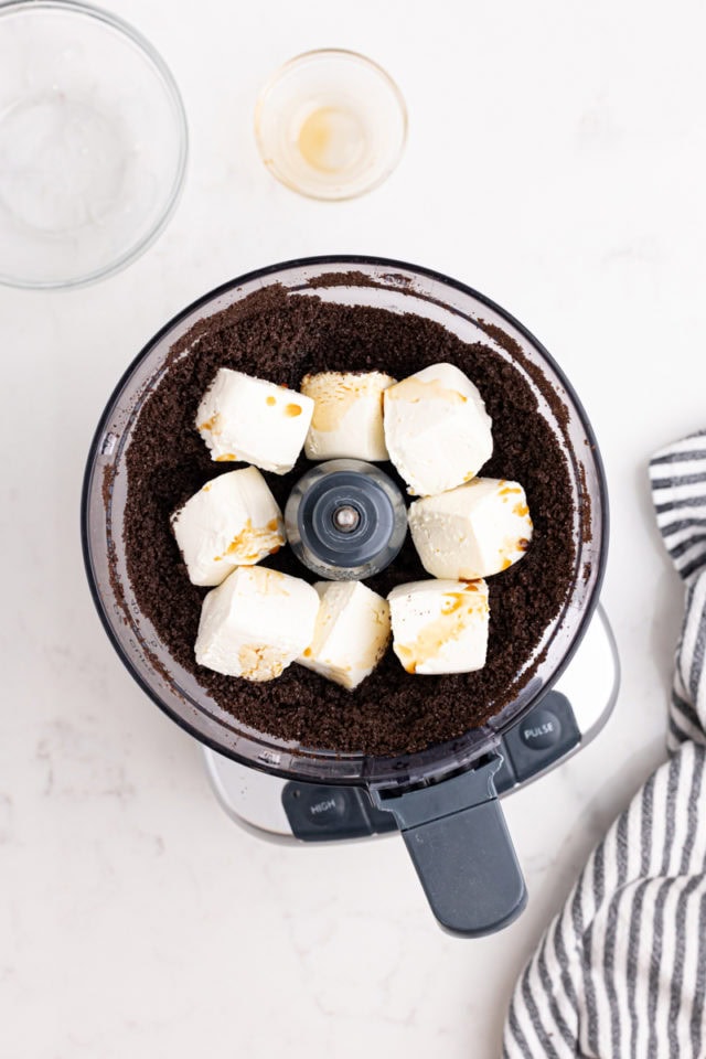 overhead view of cream cheese and vanilla added to Oreo crumbs in the bowl of a food processor