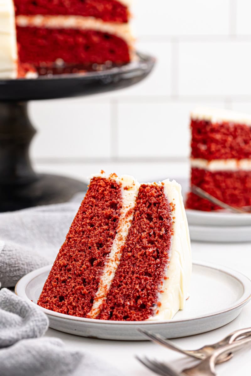 a slice of red velvet cake on a white plate with more cake in the background