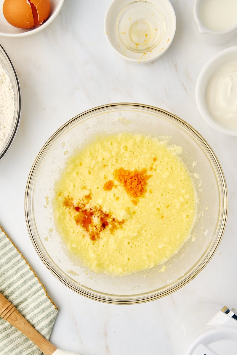 Overhead view of vanilla extract and orange zest being added to the developing cake batter in a mixing bowl.